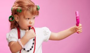 Little girl playing with mothers makeup and holding mirror on pink background